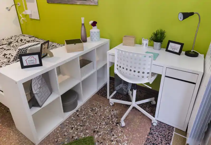 Angolo camera compatto con scaffale bianco e scrivania ordinata accanto al letto; stile moderno e luminoso con accessori decorativi e una sedia studio.