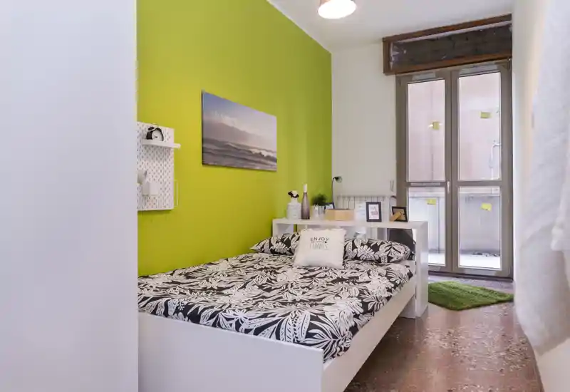 Camera da letto luminosa e ben allestita con letto matrimoniale, biancheria decorativa e parete accentuata verde, con porta finestra che porta luce naturale.