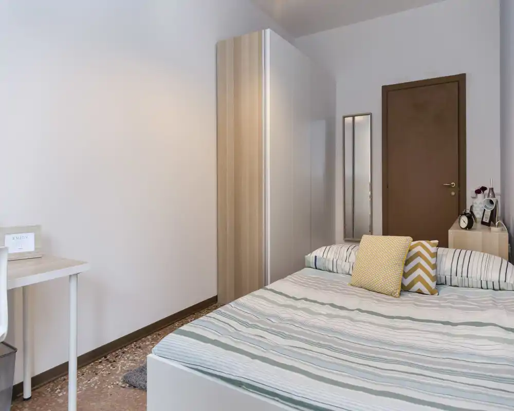 Camera da letto luminosa e ordinata con letto matrimoniale, cuscini decorativi, armadio e piccola scrivania — ideale per un annuncio in affitto che punta su comfort e spazio.
