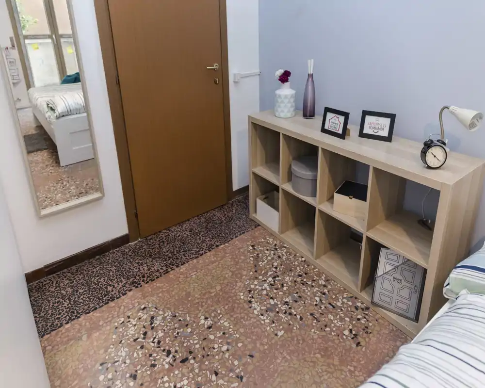 Angolo camera compatto con letto singolo parzialmente visibile sul bordo destro, una libreria bassa a cubi con oggetti decorativi e uno specchio a figura intera che riflette il resto della stanza. Lo spazio è ordinato, ben illuminato e adatto a una persona.