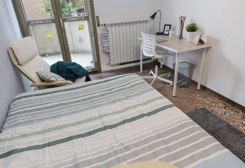 Chambre lumineuse et simplement meublée avec un grand lit, un fauteuil près de la fenêtre et un petit bureau. La lumière naturelle et les tons neutres offrent une atmosphère calme et cosy.