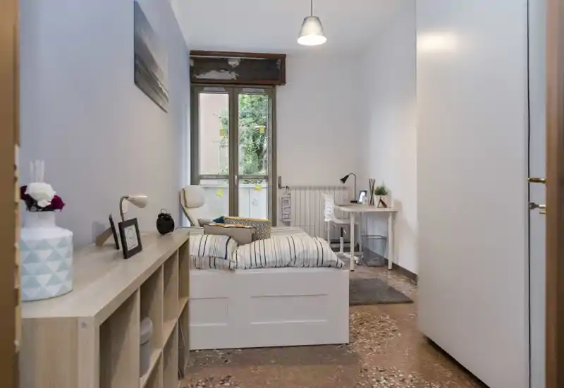 Chambre lumineuse et soignée avec un lit blanc, des textiles confortables et un petit espace de travail près de la fenêtre. La lumière naturelle et la décoration neutre donnent une ambiance calme et accueillante.