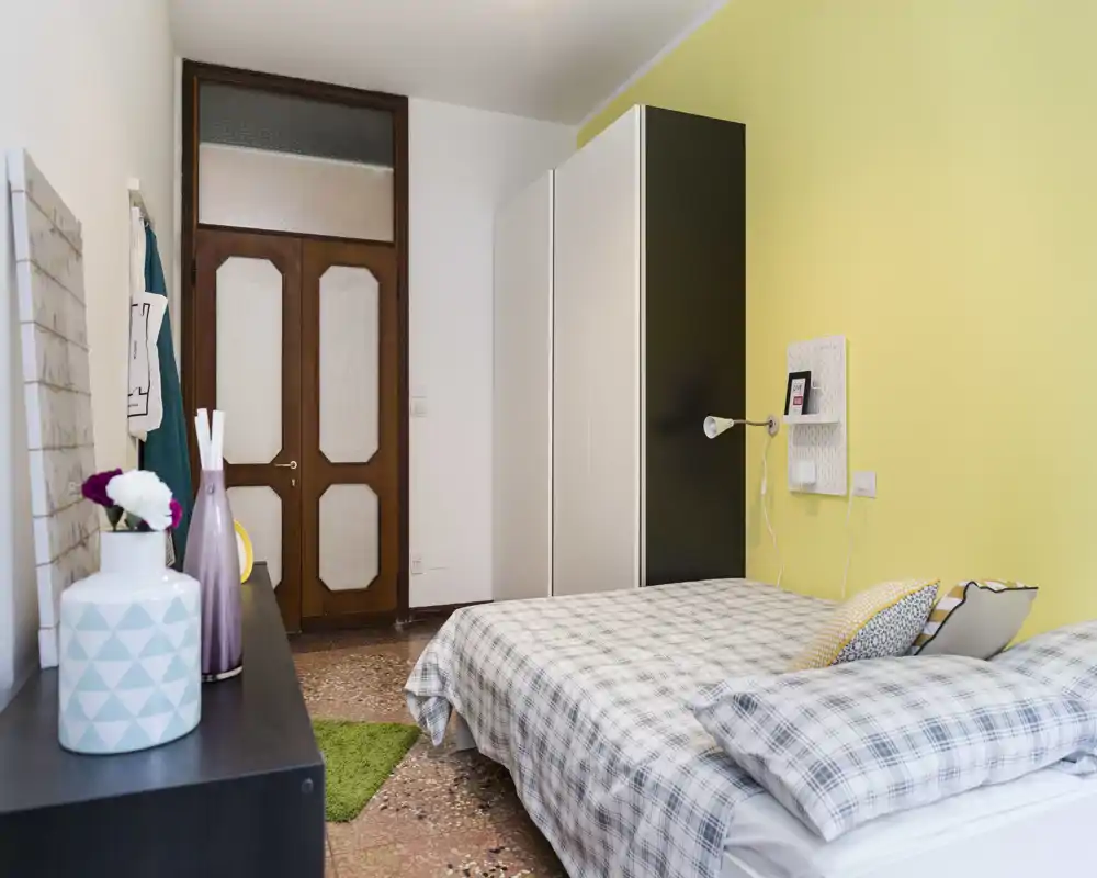 Dormitorio luminoso y ordenado con cama doble, ropa de cama a cuadros y un amplio armario frente a una pared amarilla suave, ideal para anuncios que destaquen confort y espacio de almacenamiento.