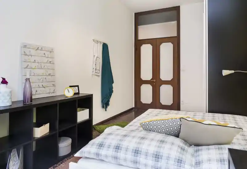 Camera da letto luminosa e ordinata con biancheria a quadretti e cuscini decorativi. Un letto basso occupa il primo piano, mentre mensole e decorazioni a parete creano un’atmosfera accogliente e moderna.