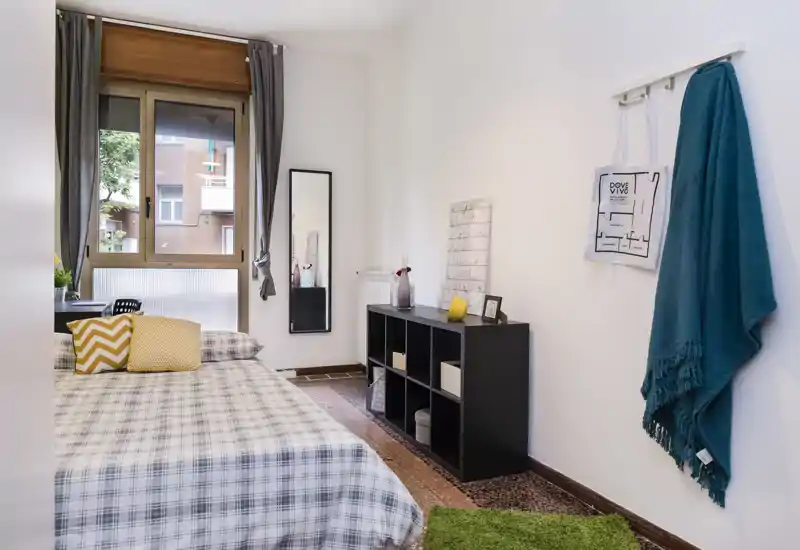 Camera da letto luminosa e moderna con grande finestra, biancheria a quadri e cuscini decorativi; mensole ordinate e ganci a muro per riporre gli oggetti.