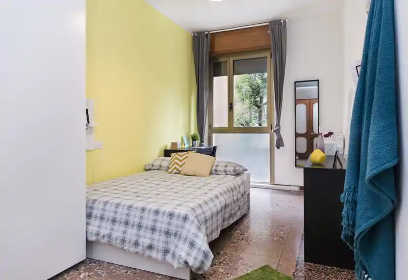 Camera da letto luminosa e ordinata con letto matrimoniale contro una parete gialla, comodino, cuscini decorativi e finestra che illumina la stanza.