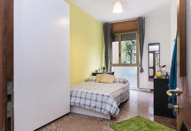 Camera da letto luminosa e accogliente con letto matrimoniale con copriletto a quadri, parete gialla a contrasto, comodino con piante e finestra che illumina la stanza.
