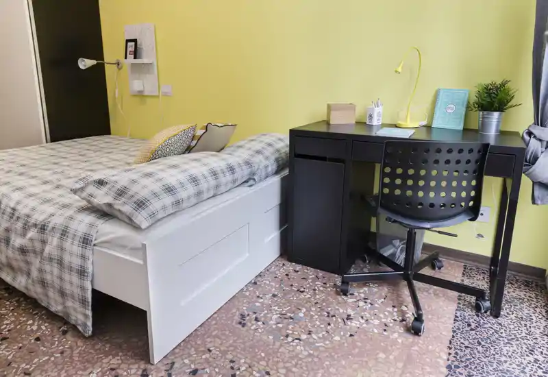 Camera da letto luminosa e accogliente con letto bianco, copriletto a quadri e una scrivania nera compatta—ideale per studenti o affitti brevi. Stile pulito, cuscini decorativi e un piccolo angolo studio rendono la stanza funzionale.
