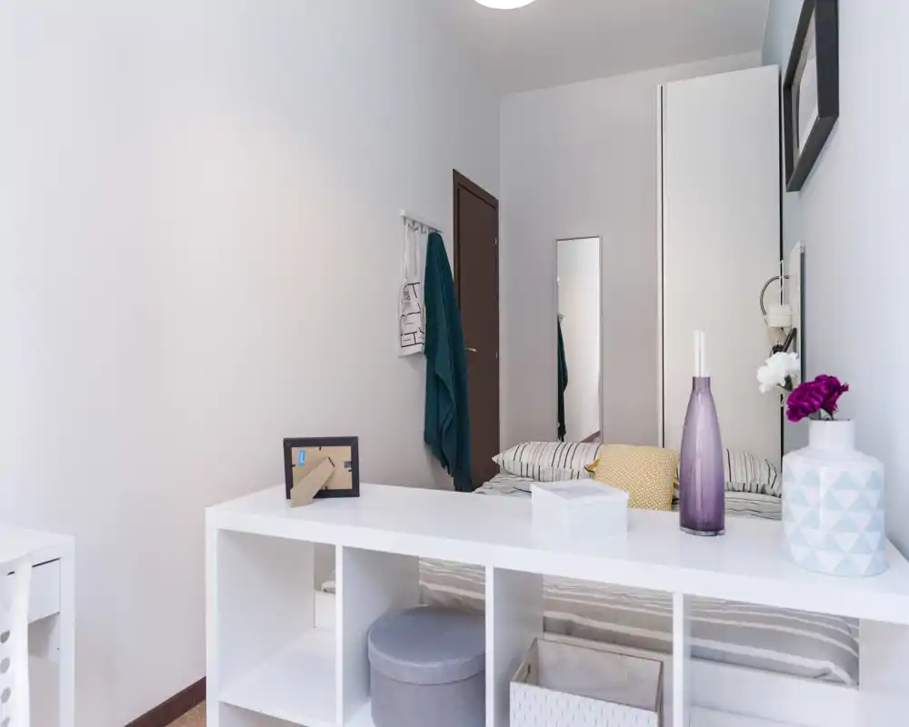 Camera da letto luminosa e ordinata con letto visibile, cuscini decorativi e un mobile a scaffali bianchi in primo piano; palette neutra e arredi minimal per un’atmosfera moderna e rilassante.