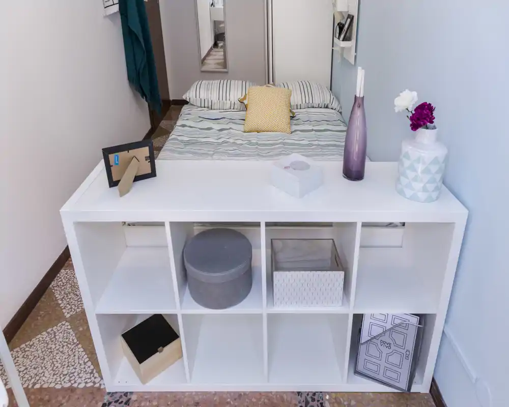 Camera da letto ordinata e luminosa con un letto matrimoniale dietro un basso scaffale bianco e dettagli decorativi morbidi per un'atmosfera accogliente e moderna.