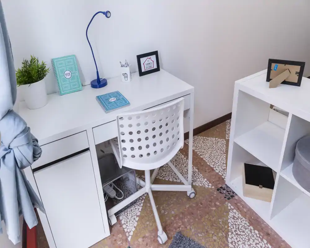 Piccolo angolo ufficio in casa, con scrivania bianca, sedia girevole forata, scaffale e accessori decorativi. Spazio funzionale e ordinato per una persona.