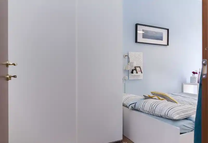 Camera da letto luminosa e accogliente con letto ben rifatto, biancheria a righe e pareti azzurre; comodino compatto e quadro incorniciato per un ambiente rilassante.