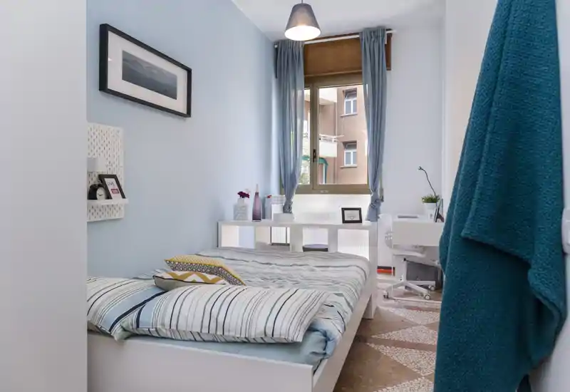 Camera singola luminosa e accogliente con letto ordinato, pareti azzurre e un angolo studio vicino alla finestra — ideale per un soggiorno confortevole.
