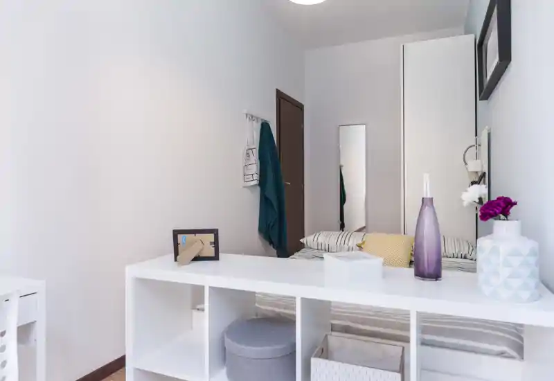 Bright, neatly staged bedroom with a visible double bed, decorative cushions and a white open-shelving unit in the foreground; soft neutral palette and minimal decor create a calm, modern atmosphere.