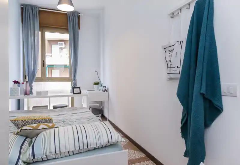 Camera da letto luminosa e ordinata con letto a righe, cuscini e una scrivania vicino alla finestra — ideale per annunci che vogliono mostrare un'area notte accogliente e ben illuminata.