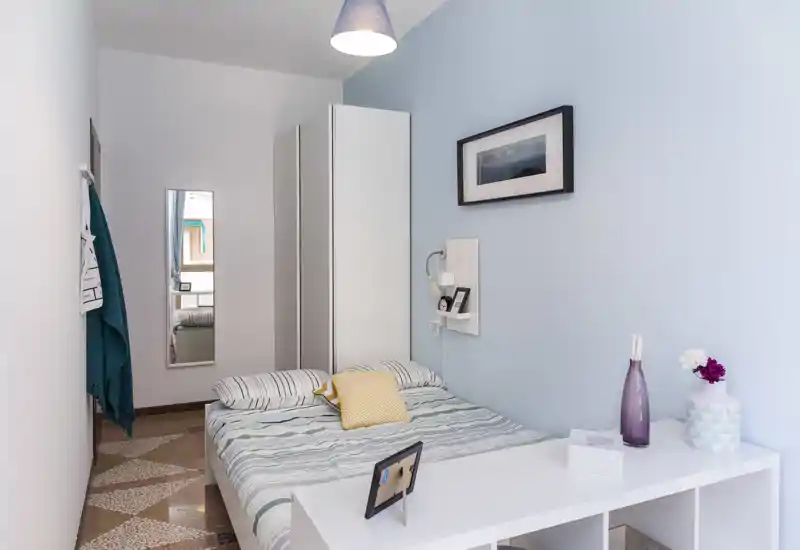 Camera da letto compatta e luminosa con letto matrimoniale, armadio e mensola scrivania; parete azzurra e tessuti accoglienti rendono l'ambiente rilassante.