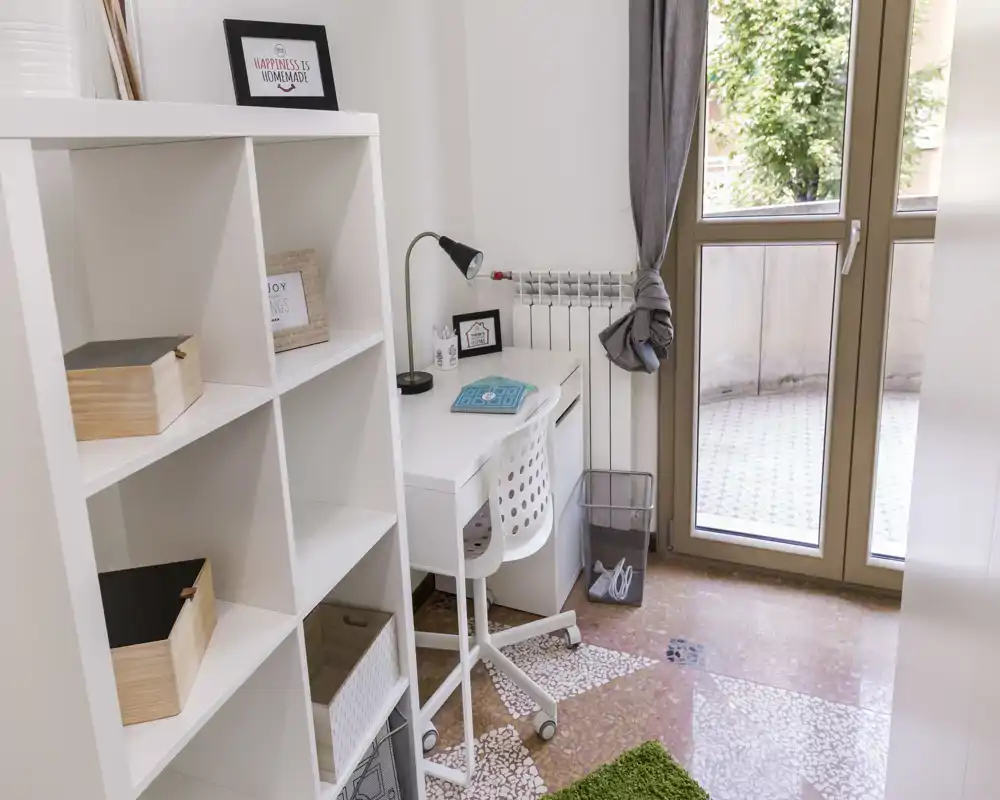 Pièce lumineuse avec une étagère blanche et un petit bureau près d'une porte-fenêtre donnant sur le balcon ; coin bureau moderne et soigné, bien éclairé.