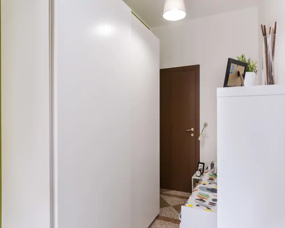 Coin de chambre avec une grande armoire blanche et une petite partie du lit visible avec une couverture à motifs ; mobilier net et porte en bois donnant un espace compact et rangé.