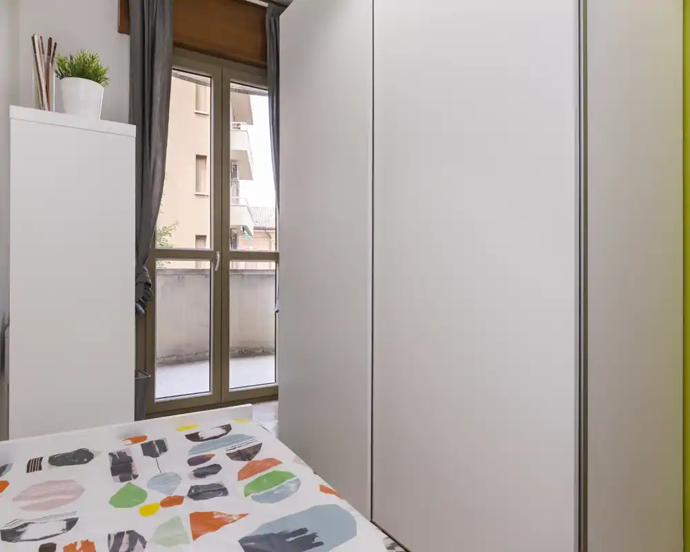 Chambre compacte avec couette à motifs, grand placard blanc et porte-fenêtre donnant sur le balcon apportant de la lumière naturelle ; espace propre, moderne et fonctionnel, adapté pour une personne ou une chambre d'appoint.