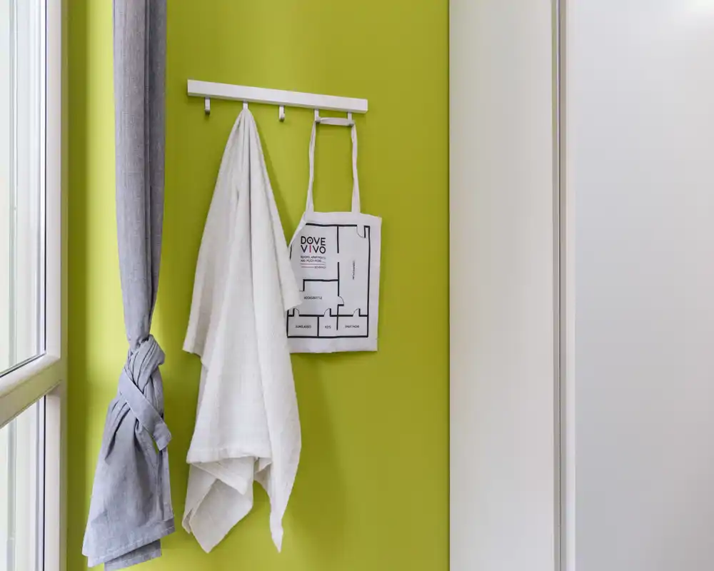 Coin d'entrée avec patères murales portant deux tissus et un tote bag, mur vert vif et fenêtre voisine apportant la lumière naturelle.