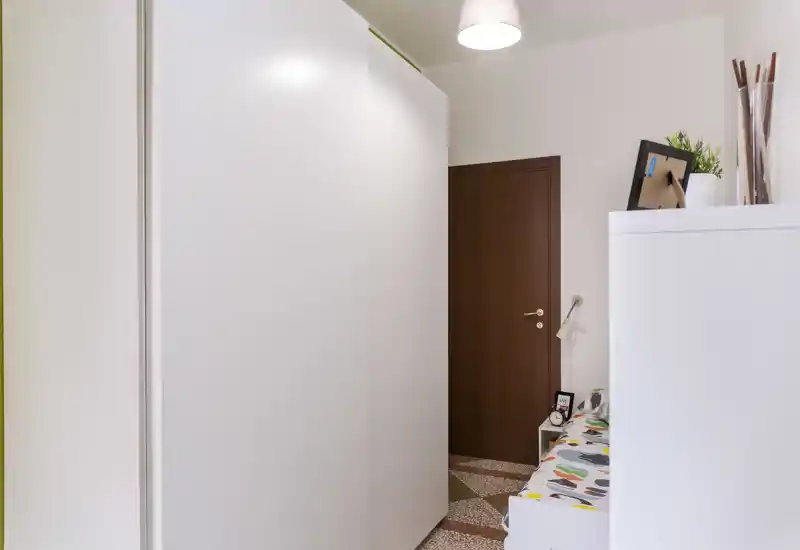 Coin de chambre avec une grande armoire blanche et une petite partie du lit visible avec une couverture à motifs ; mobilier net et porte en bois donnant un espace compact et rangé.