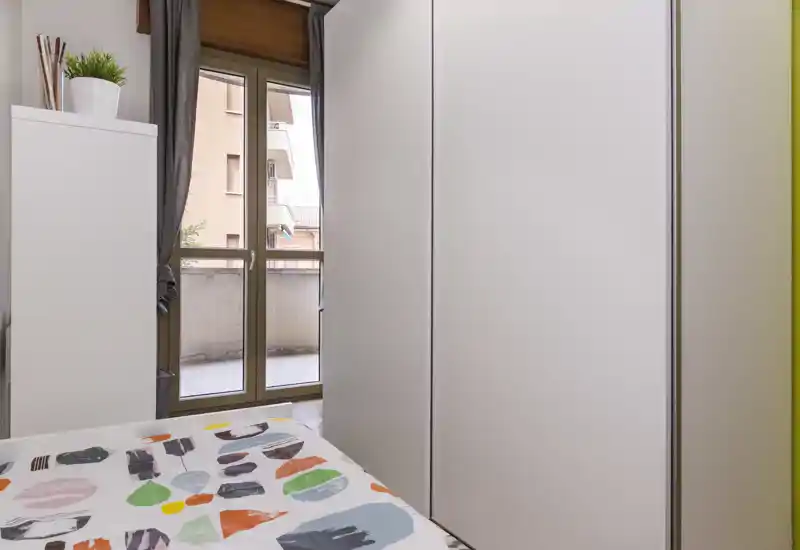 Chambre compacte avec couette à motifs, grand placard blanc et porte-fenêtre donnant sur le balcon apportant de la lumière naturelle ; espace propre, moderne et fonctionnel, adapté pour une personne ou une chambre d'appoint.