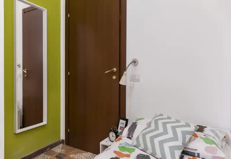 Coin de chambre avec un lit simple habillé d'une couette colorée, une table de chevet avec réveil, une lampe murale de lecture et une glace pleine longueur à côté d'une porte en bois.
