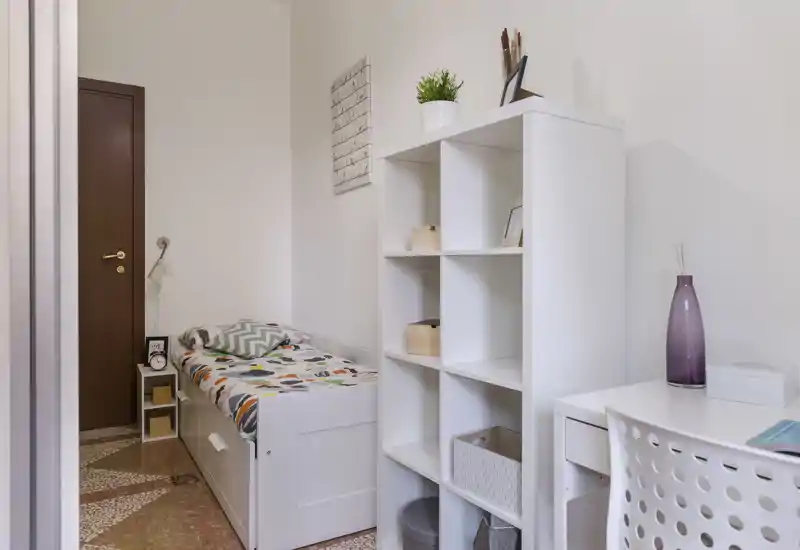 Chambre compacte et lumineuse avec un lit simple, tiroirs de rangement intégrés et une étagère blanche; un coin bureau propre et des textiles apportent une atmosphère chaleureuse.
