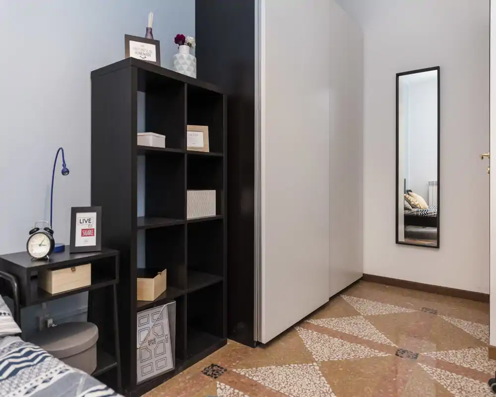 Rincón de dormitorio con mesita, estantería y parte de la cama visible; armario alto y almacenaje moderno para buena organización.