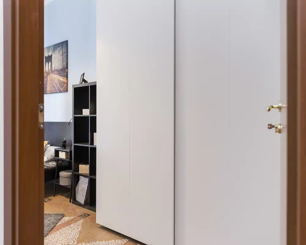 Vista de un dormitorio a través de una puerta abierta con la cama parcialmente visible, mesita y estantería; tonos neutros y decoración moderna y sencilla que transmiten funcionalidad y confort.