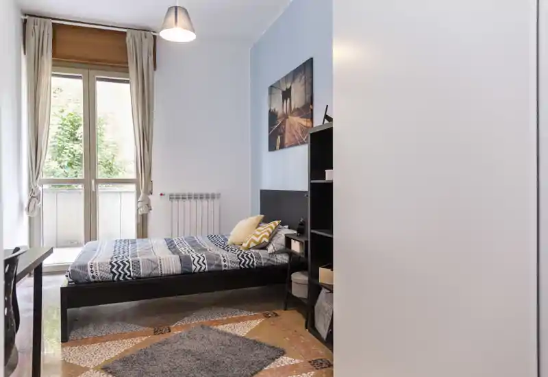 Dormitorio luminoso y ordenado con cama doble, ropa de cama estampada y cojines decorativos; gran ventana con cortinas que aporta abundante luz natural y un ambiente moderno.