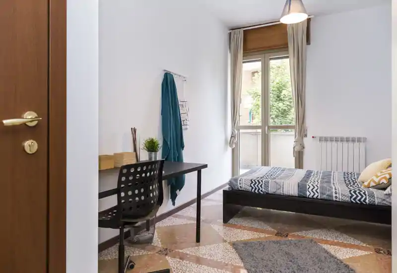 Habitación individual luminosa y ordenada con escritorio junto a la pared, ropa de cama con estampado y ventana que aporta luz natural — ideal para un estudiante o una persona sola.