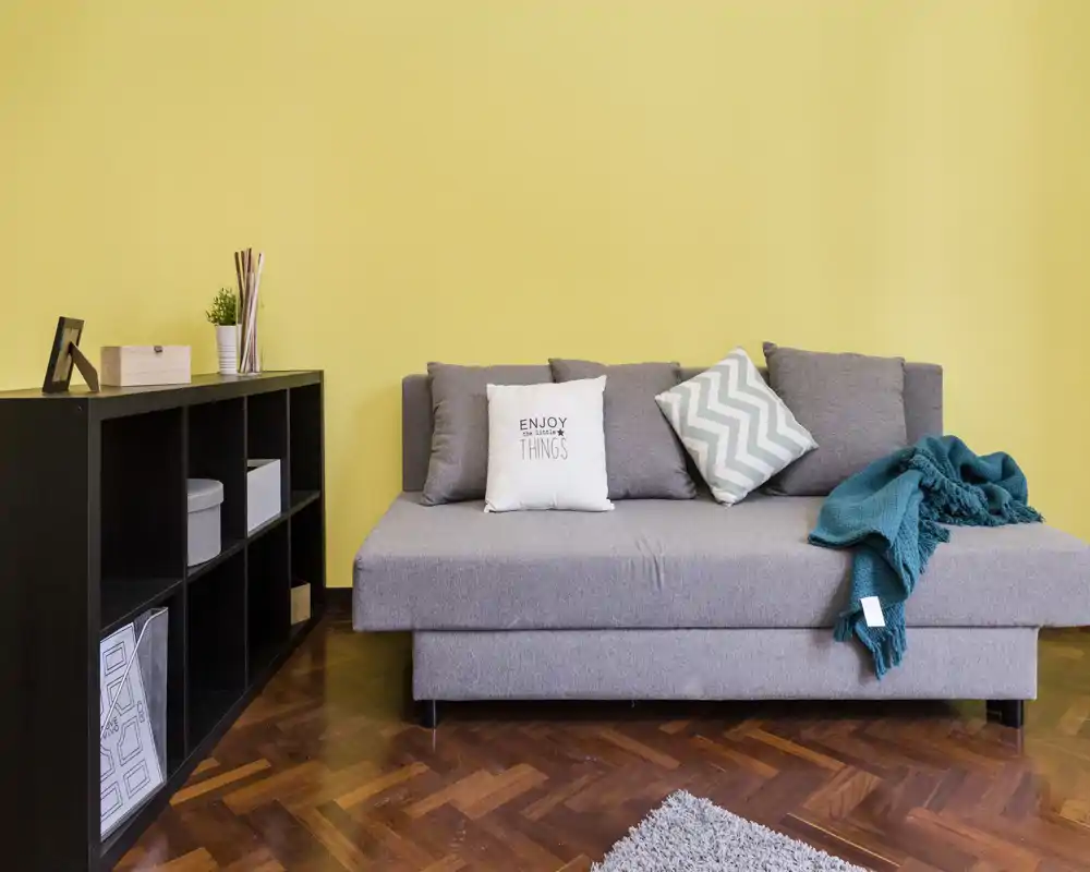 Zona giorno luminosa e moderna con divano grigio, cuscini decorativi e una coperta turchese su una parete gialla; mobile nero con vani per contenere oggetti.