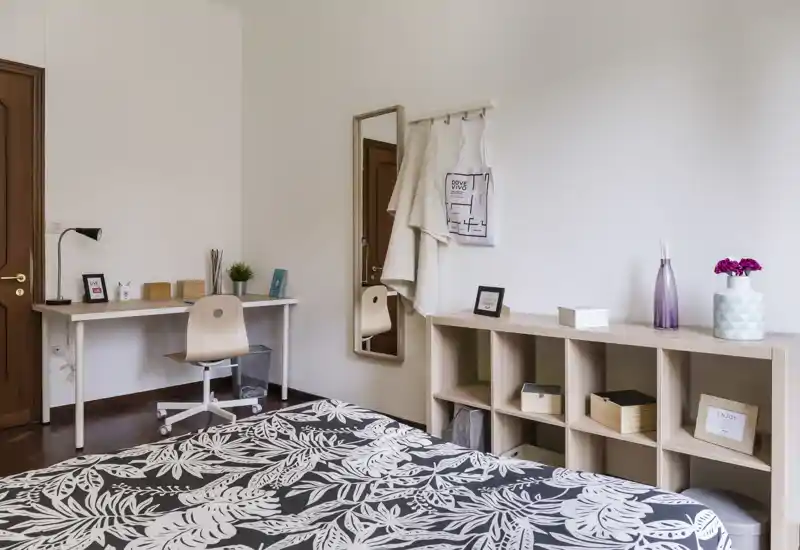 Camera da letto luminosa e moderna con una coperta a disegni in primo piano, una scrivania e scaffalatura lungo la parete e accessori decorativi semplici per un ambiente accogliente.