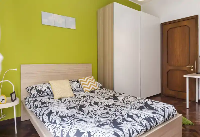 Camera da letto luminosa e moderna con letto matrimoniale, copripiumino a fantasia e parete accentata verde; armadio e comodino completano l’arredo.