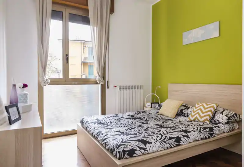 Camera da letto moderna e luminosa con letto matrimoniale, biancheria a fantasia e parete verde a contrasto; la luce naturale dalla finestra rende l’ambiente accogliente.