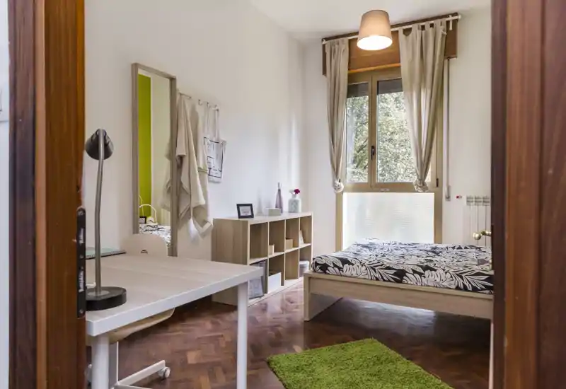 Camera da letto luminosa e ben arredata con letto matrimoniale, scrivania e ampia finestra; mobili in legno e tessili morbidi creano un ambiente accogliente.