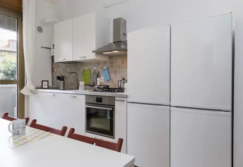 Bright, modern fitted kitchen with white cabinets, built-in oven and two tall fridges, plus a small dining table in the foreground — practical and well-lit for everyday use.