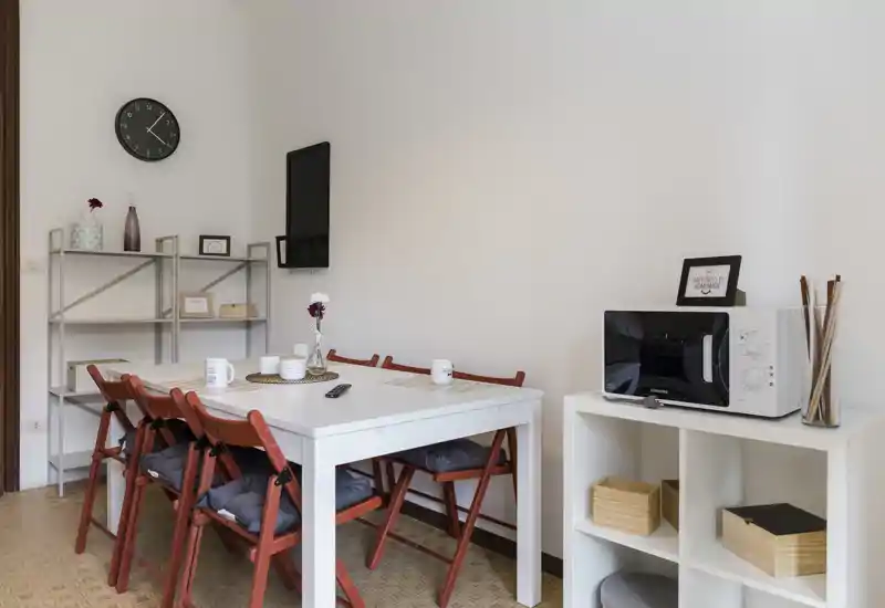 Bright, tidy dining area with a white table, four red folding chairs with cushions, simple shelving and a microwave on a cubed side unit — ideal for compact apartment listings.