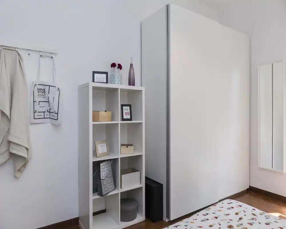 Coin de chambre petit et lumineux avec une armoire blanche et une étagère à cubes; le linge de lit à motifs est partiellement visible. Style moderne et épuré avec boîtes et vases décoratifs qui apportent une note chaleureuse.