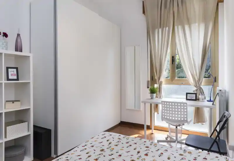 Camera da letto luminosa e ben arredata con una trapunta decorata, una scrivania compatta vicino alla finestra e un grande armadio bianco — ideale per una persona o soggiorni brevi.