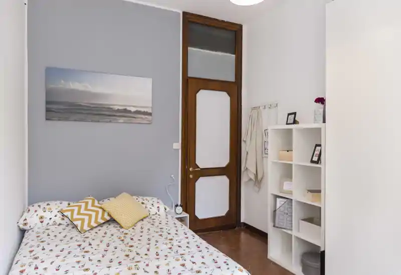 Camera da letto luminosa e ordinata con letto completo, biancheria decorata e cuscini; porta in legno e libreria offrono spazio e un'atmosfera accogliente.