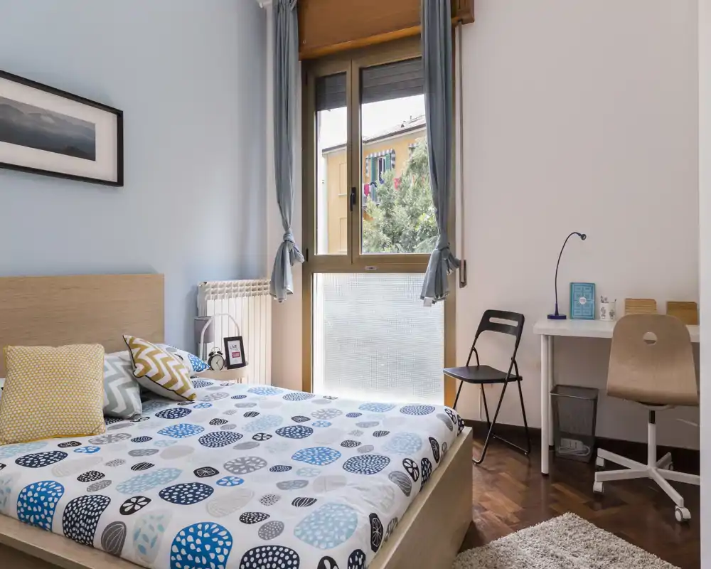 Chambre lumineuse et bien mise en scène avec un lit double, une parure de lit à motifs et une grande fenêtre apportant la lumière naturelle ; un petit bureau et des chaises offrent un espace de travail pratique.