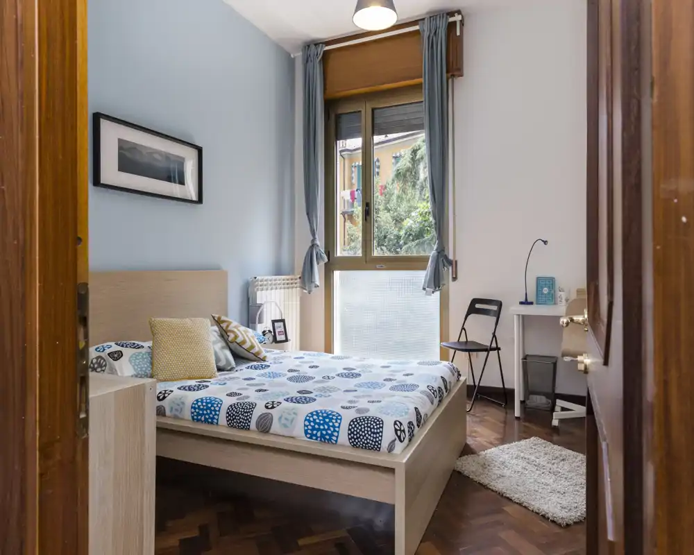 Chambre lumineuse et chaleureuse avec un lit double, une couette à motifs et des coussins moelleux. Grande fenêtre apportant de la lumière naturelle, coin bureau simple et parquet chaleureux.