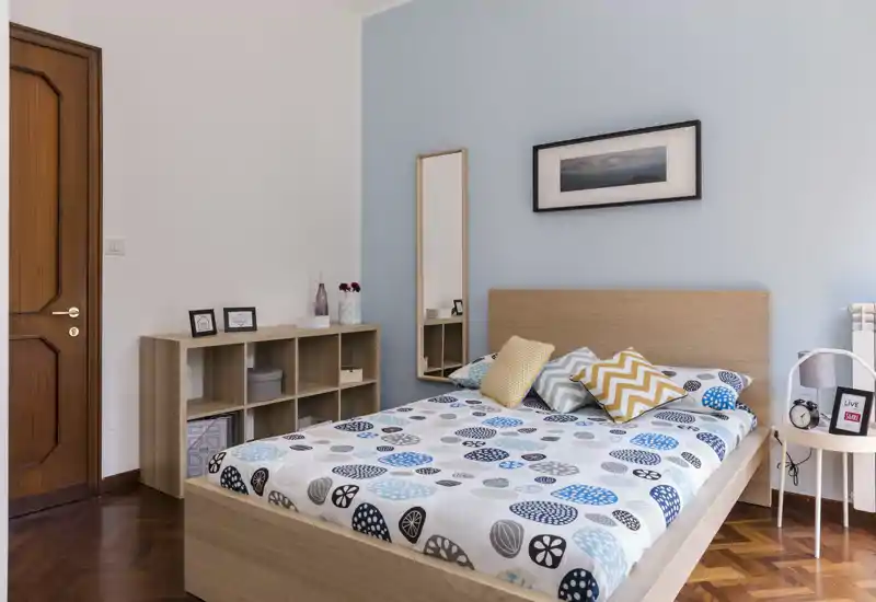 Chambre lumineuse et moderne avec lit double, couette à motifs et coussins décoratifs. Lumière naturelle, mur bleu pâle et meubles en bois clair donnent une atmosphère calme et accueillante, idéale pour une annonce.