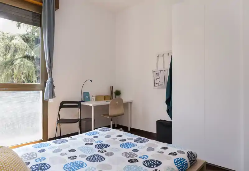 Camera luminosa e arredata in modo essenziale con copriletto a motivi, scrivania vicino alla finestra e armadio bianco — perfetta per una persona o uno studente.