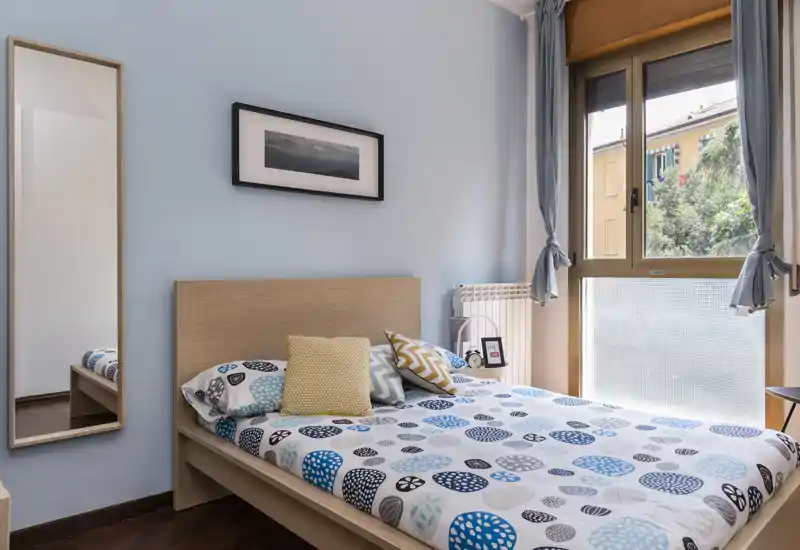 Chambre lumineuse et soignée avec lit en bois, linge de lit à motifs et coussins décoratifs ; grande fenêtre apportant de la lumière naturelle et une vue agréable.