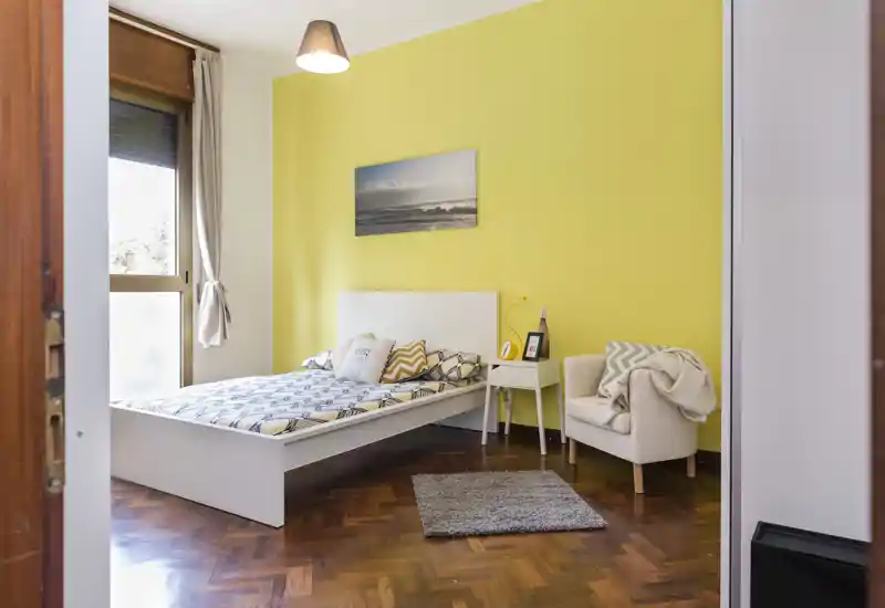 Camera da letto luminosa e ordinata con letto basso bianco, biancheria a motivi e una poltrona accogliente accanto al comodino; ampia finestra con luce naturale e parete d'accento gialla che dà carattere.