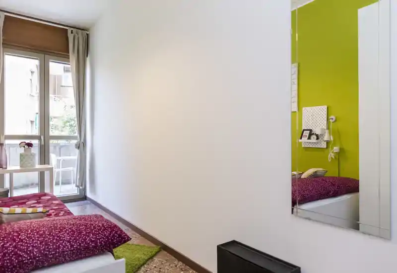 Small, bright bedroom with a single bed dressed in patterned bedding, a green accent wall reflected in a mirror, and a glass door leading to a small balcony that brings natural light.