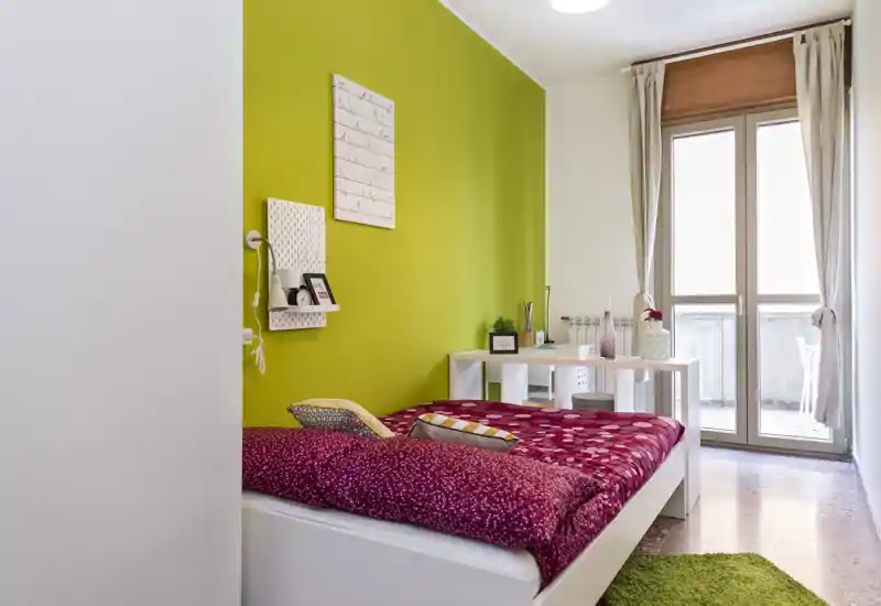 Bright, styled bedroom with a full-size bed dressed in patterned red bedding, a green accent wall and a desk by a large balcony window letting in abundant natural light.